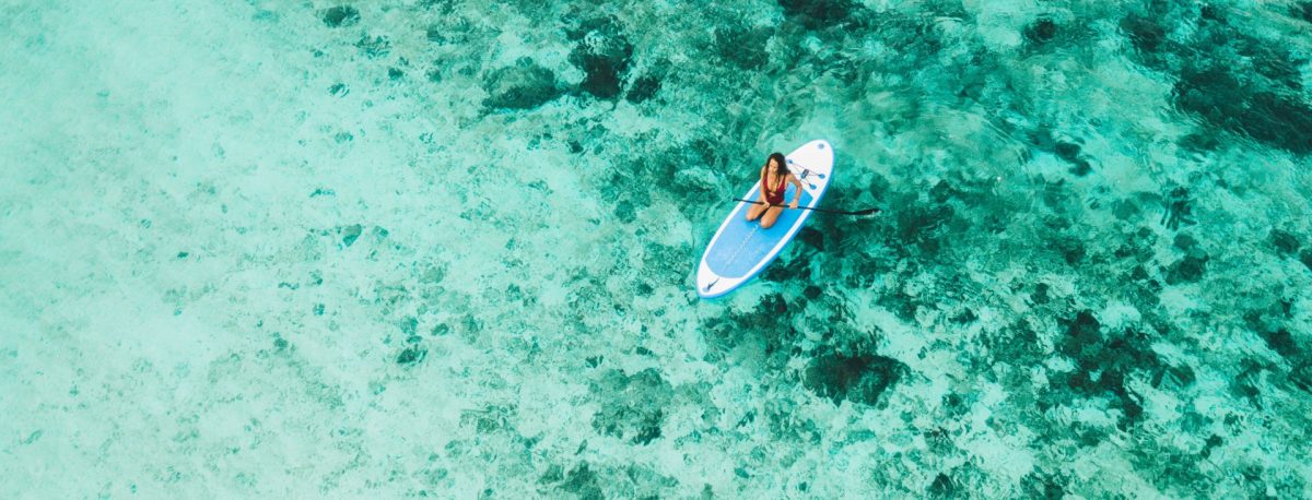 An overhead aerial view of a woman kneeling on a blue paddleboard in crystal-clear turquoise waters, showcasing the popular outdoor watersports available to visitors booking with Escape to Marco.